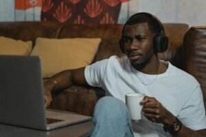 An adult man wearing headphones enjoys coffee and works on a laptop indoors, embodying remote work lifestyle.