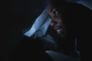 A woman smiles while using her smartphone under the covers in a dark room.