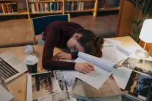 A tired woman asleep at a desk with papers in a library setting, illustrating overwork.